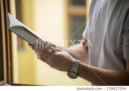 Side view of man reading book by window with warm light and shallow depth of field Side view of man reading book by window with warm light and shallow depth of field 139027504