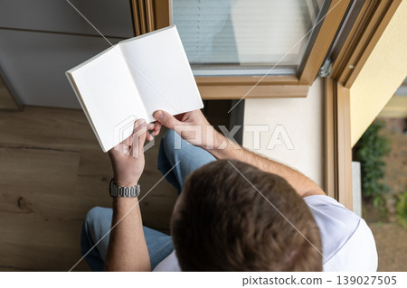 Top view of man reading book by window with natural light in cozy home interior Top view of man reading book by window with natural light in cozy home interior 139027505