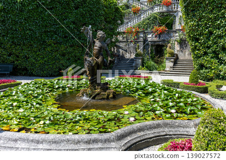 Decorative garden fountain with sculpture surrounded by flowers and classical architecture 139027572