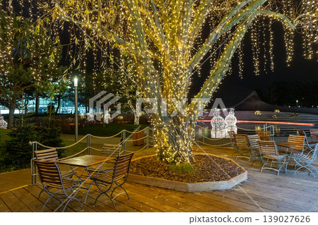 Outdoor festive lighting with illuminated tree and cozy seating area night holiday atmosphere Outdoor festive lighting with illuminated tree and cozy seating area night holiday atmosphere 139027626