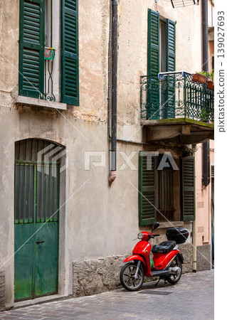 A bright red scooter parked along a narrow European street with aged plaster walls and green shutters. 139027693