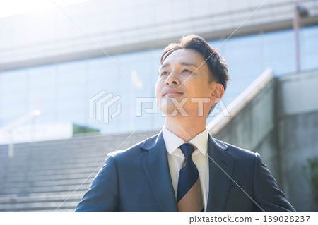 A refreshing, suit-wearing salesman, with a building in the background, looks towards the future—a business image symbolizing career success and growth. A refreshing, suit-wearing salesman, with a building in the background, looks towards the future—a business image symbolizing career success and growth. 139028237