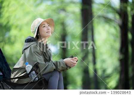 一位微笑的女子獨自坐在鬱鬱蔥蔥的森林中的露營椅上,欣賞著周圍的景色。 一位微笑的女子獨自坐在鬱鬱蔥蔥的森林中的露營椅上,欣賞著周圍的景色。 139028643