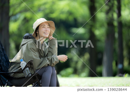A smiling woman relaxes in a camping chair amidst the lush green forest, enjoying the scenery during her solo camping trip. 139028644