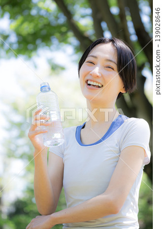 一位女性在戶外綠樹環繞中補充水分的側面肖像,她可能正在散步、跑步或其他運動中。 一位女性在戶外綠樹環繞中補充水分的側面肖像,她可能正在散步、跑步或其他運動中。 139028646
