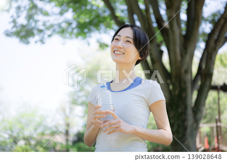一位女性在戶外綠樹環繞中補充水分的側面肖像,她可能正在散步、跑步或其他運動中鍛鍊身體。 一位女性在戶外綠樹環繞中補充水分的側面肖像,她可能正在散步、跑步或其他運動中鍛鍊身體。 139028648