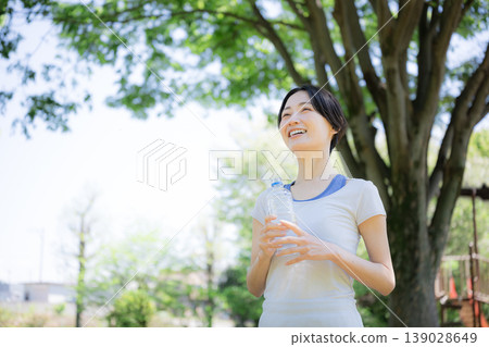 一位女性在戶外綠樹環繞中補充水分的側面肖像,她可能正在散步、跑步或其他運動中鍛鍊身體。 一位女性在戶外綠樹環繞中補充水分的側面肖像,她可能正在散步、跑步或其他運動中鍛鍊身體。 139028649