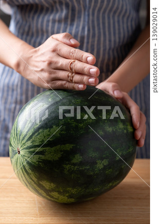 Fresh whole watermelon on a wooden cutting board with woman's hands 139029014