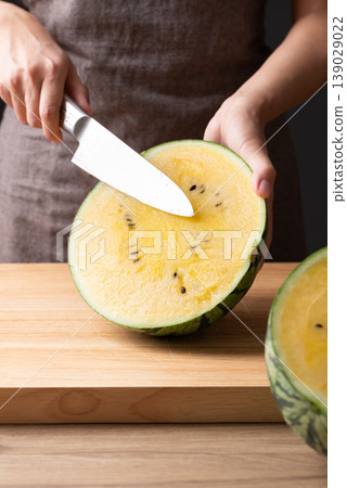 Woman's hands holding a kitchen knife to cut a yellow watermelon on a wooden cutting board Woman's hands holding a kitchen knife to cut a yellow watermelon on a wooden cutting board 139029022