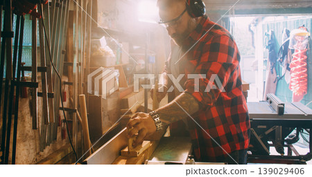 Joiner process. Furniture making. Man with ear protectors smoothing wood on thickness planer. Saw dust blur light in industry interior. Joiner process. Furniture making. Man with ear protectors smoothing wood on thickness planer. Saw dust blur light in industry interior. 139029406