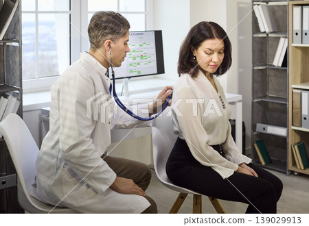 Doctor auscultating female patients back with stethoscope in bright clinic office Doctor auscultating female patients back with stethoscope in bright clinic office 139029913
