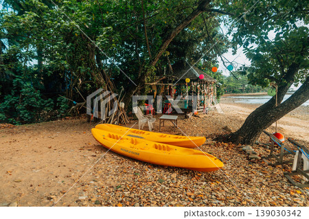 Yellow kayaks resting on a tranquil pebble beach near a rustic tropical massage hut and beach bar during a serene golden hour sunset. Yellow kayaks resting on a tranquil pebble beach near a rustic tropical massage hut and beach bar during a serene golden hour sunset. 139030342