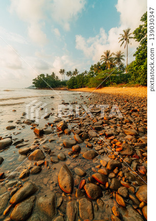Beautiful golden hour sunset reflecting off wet, smooth stones on a rocky tropical beach with a backdrop of lush green palm trees and calm ocean waves. Beautiful golden hour sunset reflecting off wet, smooth stones on a rocky tropical beach with a backdrop of lush green palm trees and calm ocean waves. 139030347