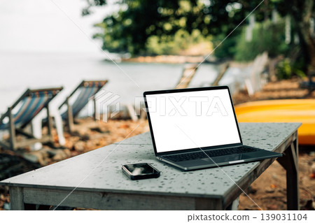 Blank white screen laptop and smartphone on a table at a tropical sandy beach. Digital nomad and remote work concept. Blank white screen laptop and smartphone on a table at a tropical sandy beach. Digital nomad and remote work concept. 139031104