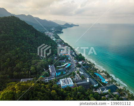 Aerial view of beachfront resorts, solar panels, and a coastal road winding through a lush tropical mountain island. Aerial view of beachfront resorts, solar panels, and a coastal road winding through a lush tropical mountain island. 139031112