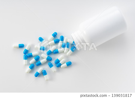 Blue and white capsules spilling from a white bottle; overhead view image against a white background. Blue and white capsules spilling from a white bottle; overhead view image against a white background. 139031817