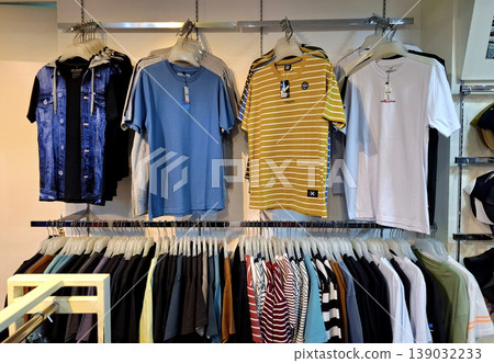 Yogyakarta, Indonesia - March 18th 2026: Rows of colorful T-shirts, a blue denim jacket, and various casual garments neatly arranged on metal hangers in a modern clothing store. Yogyakarta, Indonesia - March 18th 2026: Rows of colorful T-shirts, a blue denim jacket, and various casual garments neatly arranged on metal hangers in a modern clothing store. 139032233
