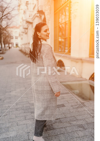 Woman coat sunset stroll. Stylish young woman in a plaid wool coat walks along an urban city street during a warm golden hour evening 139033039
