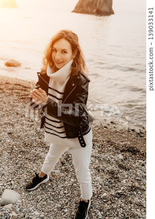 Woman beach fashion smiling lady in leather jacket and striped sweater posing on pebble seashore at sunset 139033141