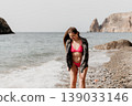 Woman beach bikini stands smiling on a pebble shore as waves crash during a summer coastal vacation trip with cliffs 139033146