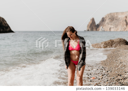 Woman beach bikini stands smiling on a pebble shore as waves crash during a summer coastal vacation trip with cliffs 139033146