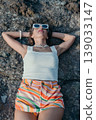 Woman beach vacation relaxing on rocky ground wearing white sunglasses and striped shorts during summer travel 139033147