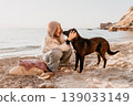 Woman dog beach petting dark mutt on a sandy pebble coastline during a tranquil golden hour. 139033149