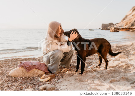 Woman dog beach petting dark mutt on a sandy pebble coastline during a tranquil golden hour. Woman dog beach petting dark mutt on a sandy pebble coastline during a tranquil golden hour. 139033149