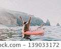 Woman, Floating, Sea enjoying summer vacation on an inflatable donut with arms raised, cliffs in the background 139033172