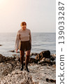Woman sunset seashore walks along a rocky coast at golden hour wearing a cozy sweater and boots 139033287