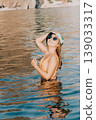 Woman Sunglasses Beach Water A woman in sunglasses and a hat stands in shallow water on a beach. 139033317