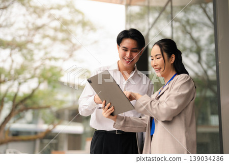 Asian businesswoman discussing report with man colleague, consultant reading paper explaining business plan to colleague, talking outside office. Asian businesswoman discussing report with man colleague, consultant reading paper explaining business plan to colleague, talking outside office. 139034286