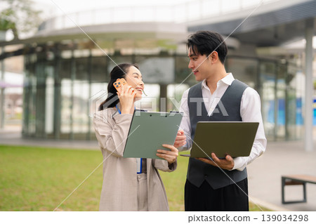 Asian businesswoman discussing report with man colleague, consultant reading paper explaining business plan to colleague, talking outside office. Asian businesswoman discussing report with man colleague, consultant reading paper explaining business plan to colleague, talking outside office. 139034298