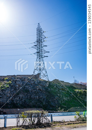 High voltage tower on sunny day near rocky terrain and road High voltage tower on sunny day near rocky terrain and road 139034545
