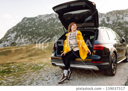 A cheerful traveler enjoying a scenic mountain view while sitting on the trunk. Travelling by car. 139037317