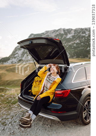 A cheerful traveler enjoying a scenic mountain view while sitting on the trunk. Travelling by car. A cheerful traveler enjoying a scenic mountain view while sitting on the trunk. Travelling by car. 139037318