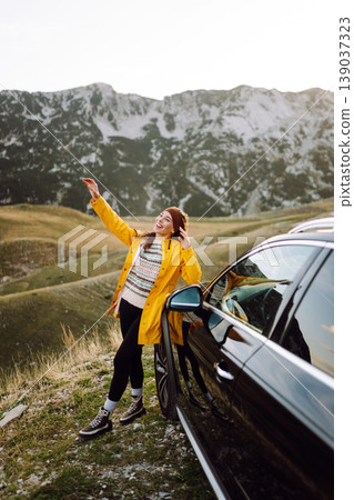 A cheerful traveler enjoying a scenic mountain view while sitting on the trunk. Travelling by car. A cheerful traveler enjoying a scenic mountain view while sitting on the trunk. Travelling by car. 139037323