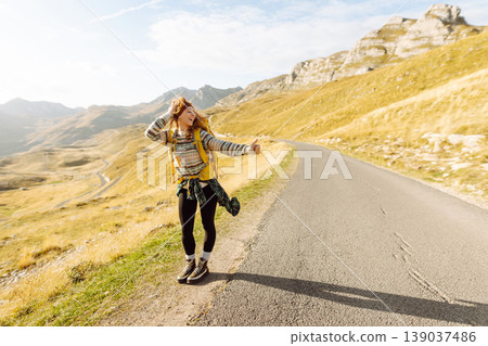 Hitchhiking woman traveler try to stop car on the mountain road. Travel and vacation theme 139037486