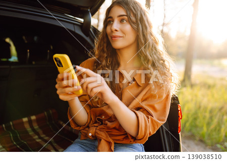 Young curly woman in stylish clothes in the trunk of a car in the forest and uses the phone. 139038510