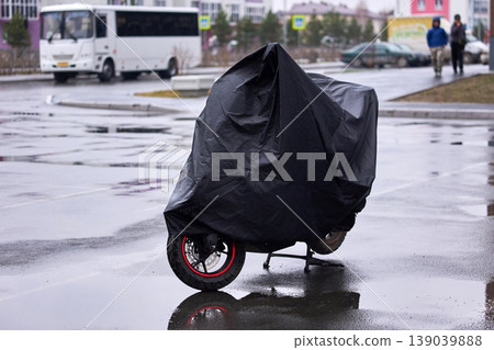 A motorcycle is covered with a black tarp and is parked on a wet street. A motorcycle is covered with a black tarp and is parked on a wet street. 139039888