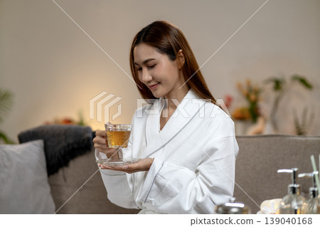 Asian adult woman enjoying warm herbal tea in white bathrobe at modern spa hotel resort room for tranquil self care experience Asian adult woman enjoying warm herbal tea in white bathrobe at modern spa hotel resort room for tranquil self care experience 139040168