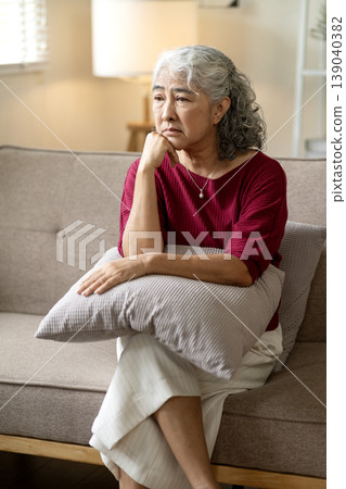Older Asian woman sit sofa deep thought worry sadness contemplating life problems mental health challenges lonely retirement senior distress contemplation solitude 139040382