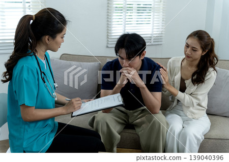 Asian medical professional offers compassionate guidance to distressed male patient alongside supportive partner during private health consultation session at home 139040396