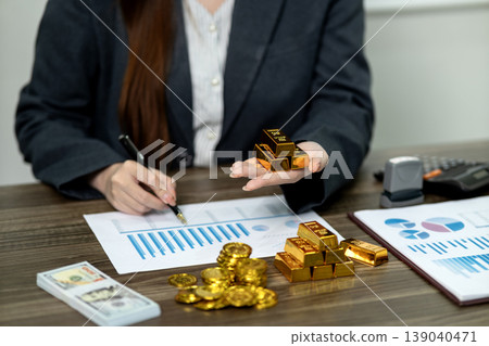 Adult Asian woman business professional analyzes financial data with pen holding gold bars and coins demonstrating wealth investment strategy office environment Adult Asian woman business professional analyzes financial data with pen holding gold bars and coins demonstrating wealth investment strategy office environment 139040471