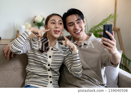 Asian young adult couple taking happy funny selfie smartphone social media casual home living room relaxing playful joyful digital lifestyle content sharing together Asian young adult couple taking happy funny selfie smartphone social media casual home living room relaxing playful joyful digital lifestyle content sharing together 139040506