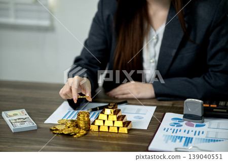 Asian adult woman investor manages financial wealth gold coins bars cash for business strategy and growth at office desk Asian adult woman investor manages financial wealth gold coins bars cash for business strategy and growth at office desk 139040551