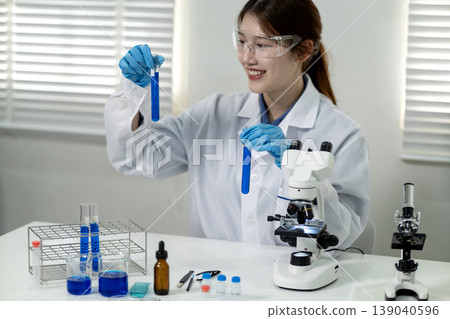 Asian adult female scientist researcher conducts chemical experiments holding test tubes blue liquid using microscope modern laboratory for scientific discovery 139040596