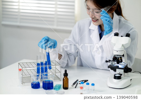 Asian adult female scientist researcher conducts experiment analysis chemical reaction laboratory setting using test tubes microscope for scientific discovery 139040598