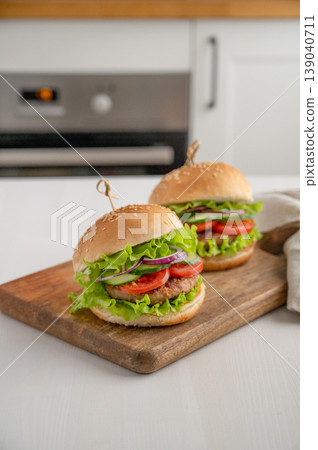 Two beef burgers with tomato, cheese, and lettuce on a cutting board on a light table Two beef burgers with tomato, cheese, and lettuce on a cutting board on a light table 139040711