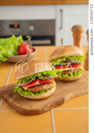 Two beef burgers with tomato, cheese, and lettuce on a cutting board on a kitchen table Two beef burgers with tomato, cheese, and lettuce on a cutting board on a kitchen table 139040713